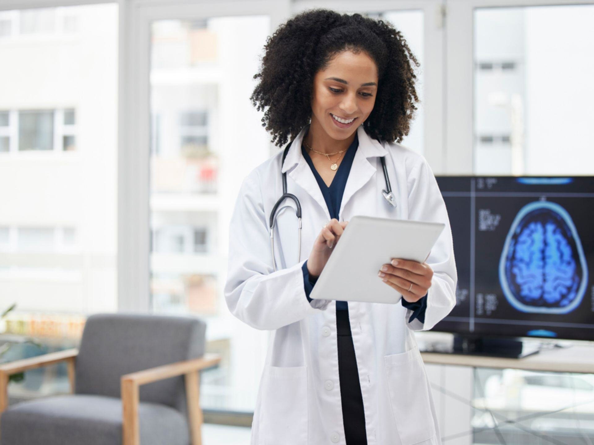 A medical professional in a white lab coat holds a tablet, with a brain scan displayed on a screen behind them. Bright, modern office setting.