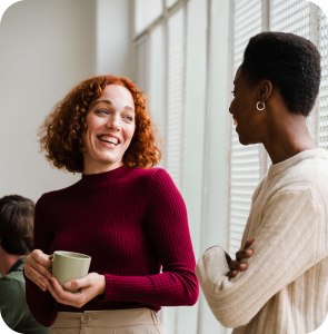 Two women smiling and conversing in an office environment, showcasing a friendly and collaborative atmosphere.